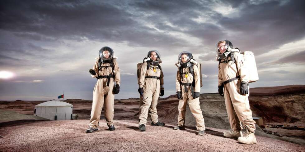 Fire av besetningsmedlemmene på Mars Desert Research Station med romdrakt på vandring utenfor basen, som sees bak. Fire av besetningsmedlemmene på Mars Desert Research Station med romdrakt på vandring utenfor basen, som sees bak.