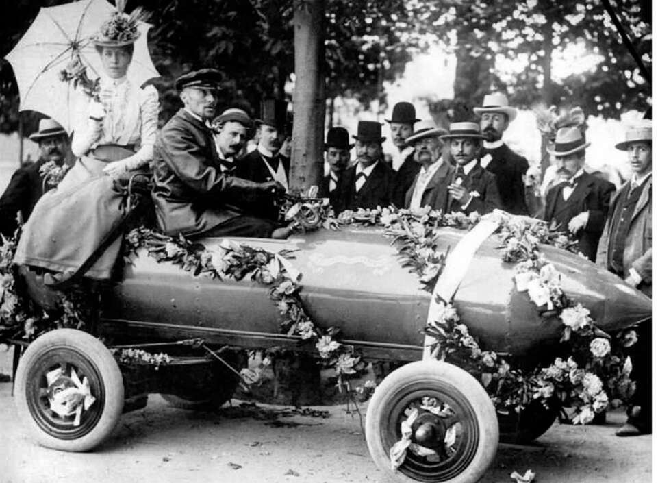 Triumfferden til Camille Jenatzy etter at han hadde sust av gårde i sin batteridrevne Jamais Contente, med en snittfart på 105 km/t over en kilometer med flygende start. Damen med paraplyen er kona hans. Jenatzy ble kalt «Den røde djevel» på grunn av hårfargen. Bilen var gråblå. Triumfferden til Camille Jenatzy etter at han hadde sust av gårde i sin batteridrevne Jamais Contente, med en snittfart på 105 km/t over en kilometer med flygende start. Damen med paraplyen er kona hans. Jenatzy ble kalt «Den røde djevel» på grunn av hårfargen. Bilen var gråblå.