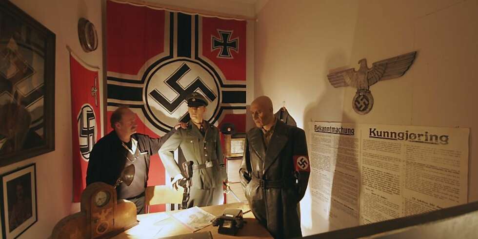 NATURTRO:Det er noe skummelt over det naturtro Gestaporommet med det svære naziflagget, nazisymbolet og plakaten med sin Bekanntmachung. Du føler at her har dødsdommene sittet løst. En av personene på bildet hører ikke naturlig hjemme i dette tidsbildet. Ser du hvem? NATURTRO:Det er noe skummelt over det naturtro Gestaporommet med det svære naziflagget, nazisymbolet og plakaten med sin Bekanntmachung. Du føler at her har dødsdommene sittet løst. En av personene på bildet hører ikke naturlig hjemme i dette tidsbildet. Ser du hvem?