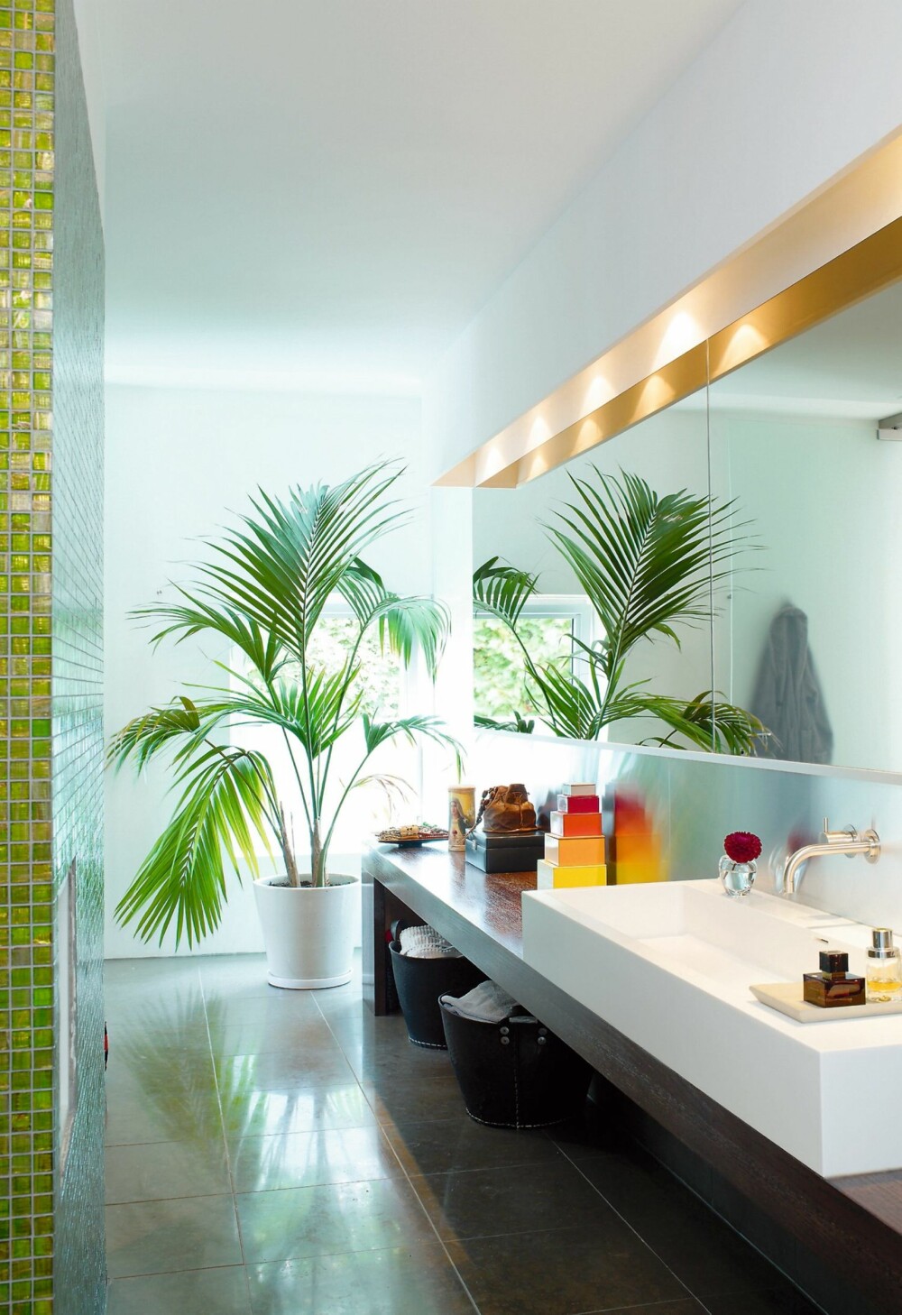 GRØNN LUNGE. Grønne planter trives på badet. og de er dekorative innslag. På gulvet er det lagt store fliser. Dette badet er stort, men smalt. Speilet på den ene siden gjør at det virker bredere.
