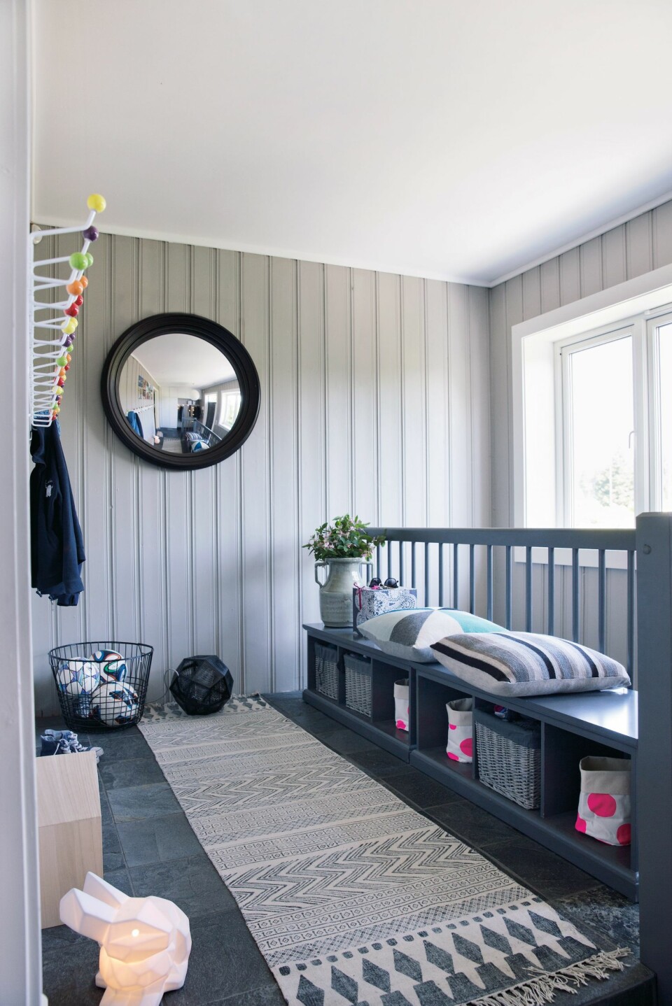 Fast plass. Smarte løsninger og gode materialvalg gjør barnefamiliens gang både åpen og bruksvennlig. Inngangspartiet har tre nivåer, øverste nivå er forbeholdt de tre aktive barna. Alt har sin plass. Under benken har de sine personlige kurver, og på veggen har de sine egne knagger. Benkene ble funnet på Skeidar. Veggfargen er Jotun Lady 10341 Kalk, mens gelenderet er malt med NCS-kode S-5000-N. Gulvlampe fra Tom Dixon, speil fra Eske, teppe og tøykurver med prikker fra Bolina. Puter og kaninlampe fra Eske. Styling Tone Kroken.