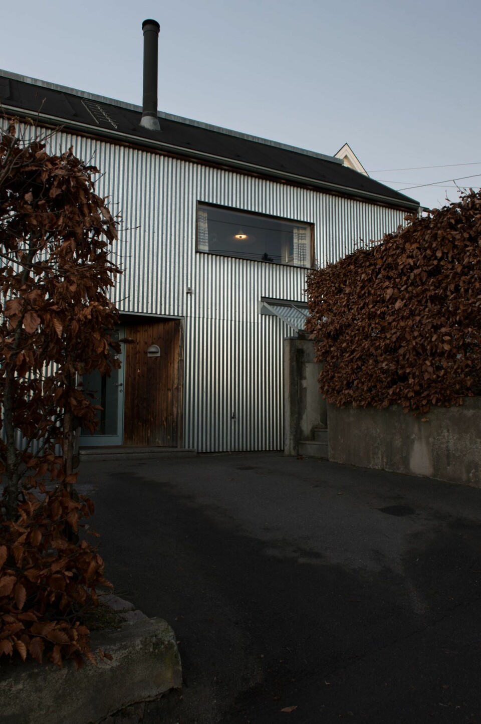 UFORGLEMMELIG: Nisjen som utgjør inngangspartiet er kledd med stående panel. Vedstabelen under overbygget til høyre bidrar ytterligere til å balansere preget av metall, betong og asfalt.