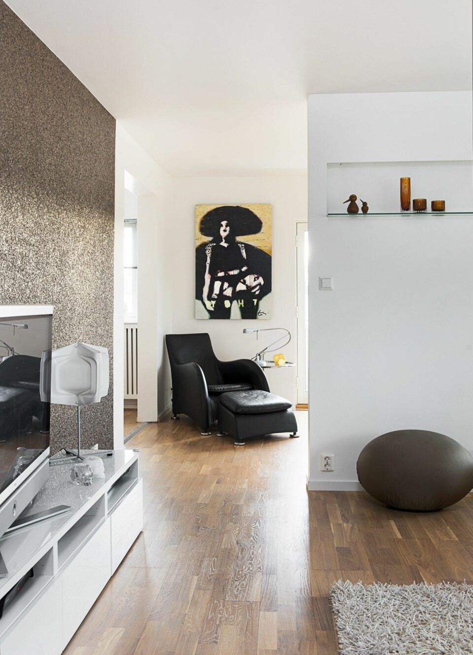 SKILLEVEGG: En lettvegg ble satt opp midt i stuen. Den har åpning på begge sider slik at det fremdeles kommer lys inn fra vinduene. Interiørarkitekten tegnet en hylle innebygget i skilleveggen, der får fine ting stå fremme. Det glitrende tapetet AKA 3004 er fra Omexco. Stolen Montis, designet av Gerard van den Berg, står i avslapningskroken, bildet er av Espen Eiborg. Bak lettveggen har beboerne etablert trenings- og arbeidsplass. Rund puff fra Cerutti Balleri.
