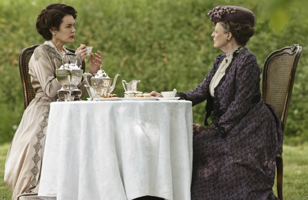 Ingenting å si på stilen når Cora drikker te med svigermor Violet ute i det fri. Tesettet er i sølv og tekoppene i fineste porselen. Elegante rottingstoler sørger for komfort og bordet er dekket med damaskduk.