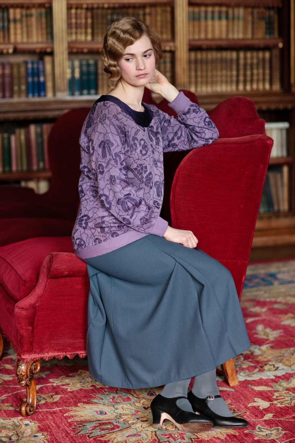 Dyp rød sofa og persisk teppe på gulvet i biblioteket på Downton Abbey. Lady Rose (Lily James) later til å kjede seg, bøkene til tross.