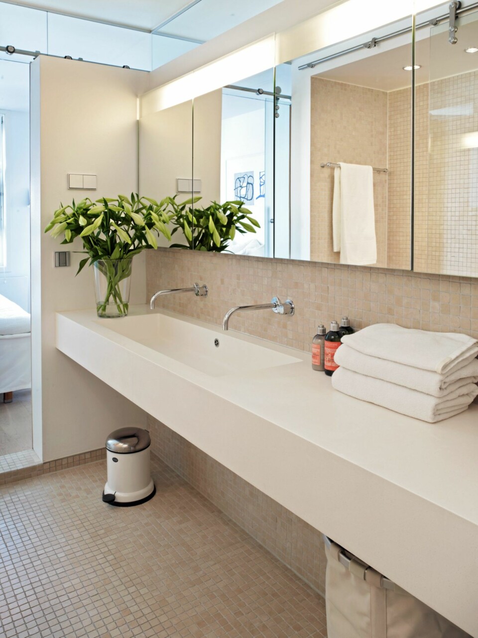 BAD I BOKS. Baderomsinnredningen består av en lang benk, med dobbel vask. Under benken er det helt åpent, og her er det derfor plass til både søppelbøtte og skittentøyskurv. Skapplass finner du derimot bak speilene, som egentlig er skapdører. Gulvet og halve veggen er flisebelagt med beige mosaikk.