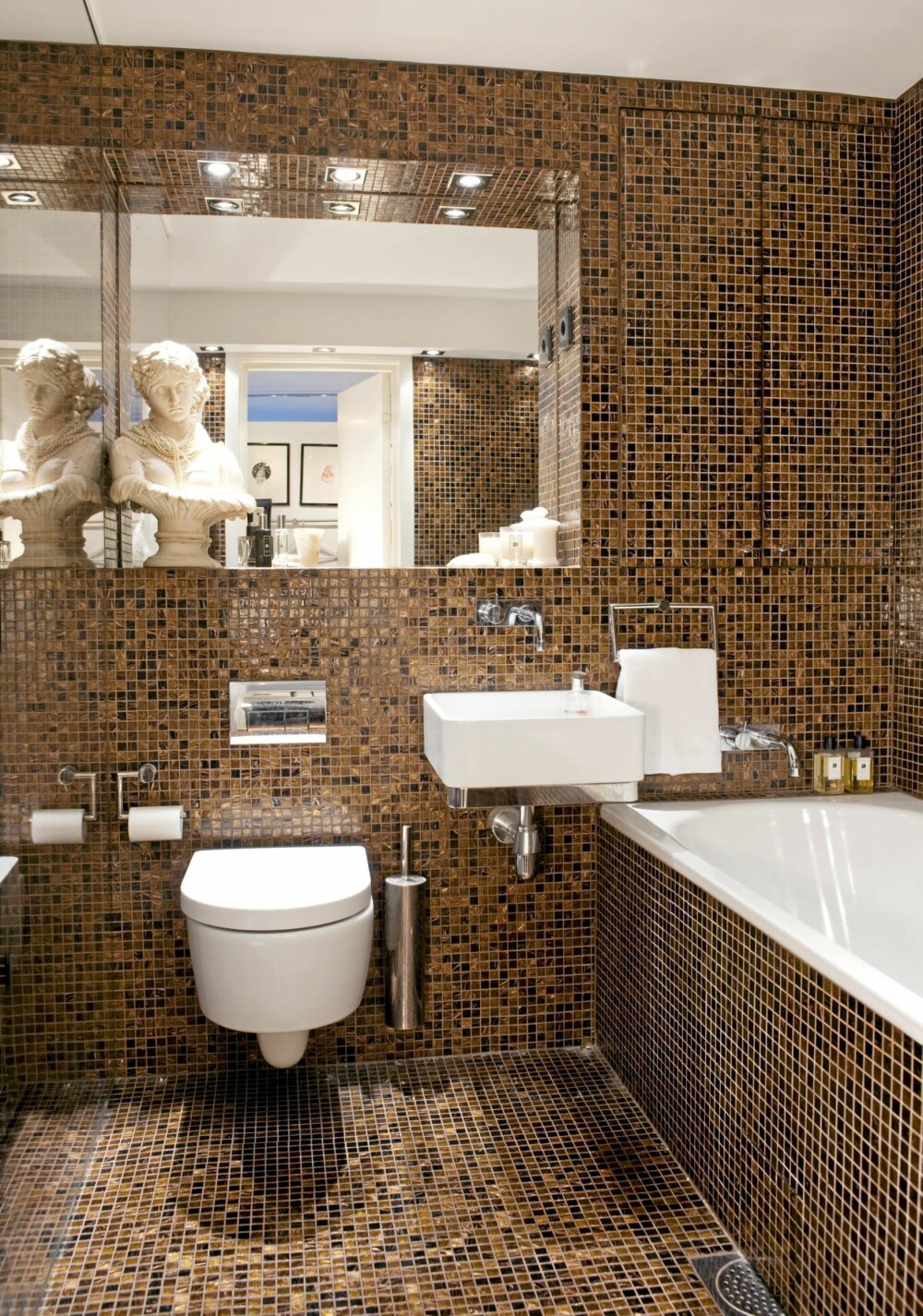 HULEFØLELSE. Hele baderommet er flisebelagt med brun mosaikk med gull fra Flisekompaniet. Det innebygde skapet ved siden av speilet er kamuflert av fliser. Speilet er som et vindu i rommet.