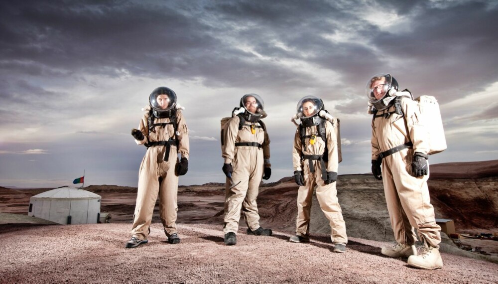 Fire av besetningsmedlemmene på Mars Desert Research Station med romdrakt på vandring utenfor basen, som sees bak.
