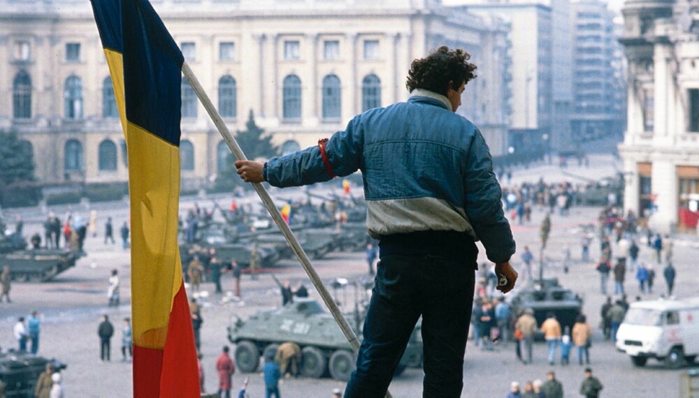 En opprører holder et rumensk flagg med kommunistemblemet fjernet mens han skuer ut over torget i Bukuresti sentrum. Stridsvognene tilhører militærenheter som har gått over til opprørernes side.