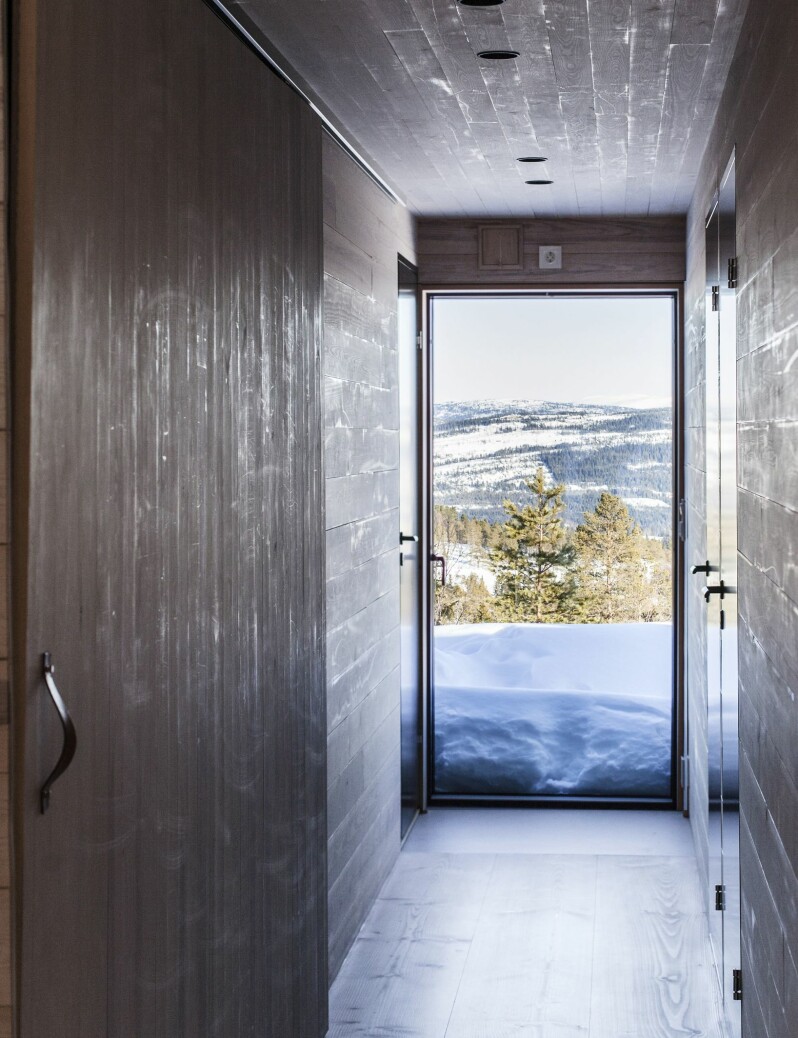 Den smale gangen utnytter åpenheten og lyset glassdøren i enden gir. Bredden på gangen er beregnet nøyaktig for å gi plass til tre, 35 cm brede, Dinesen-planker. Soverommene ligger på høyre side. Skyvedørsgarderoben til venstre ble til ved å ta litt plass fra badet, døren er laget av finerplate og kvistfri furupanel. Rett utenfor døren i enden av gangen, ved døren inn til bad og badstue, valgte paret å legge en hel flis med gulvvarme slik at man kan gå rett fra badstue og ut i snøen uten å tråkke på tregulvet. Håndtaket på skyvedøren kom Roar over på nett. De lages av en liten butikk i New York som heter Studio Zung, zung.nyc.