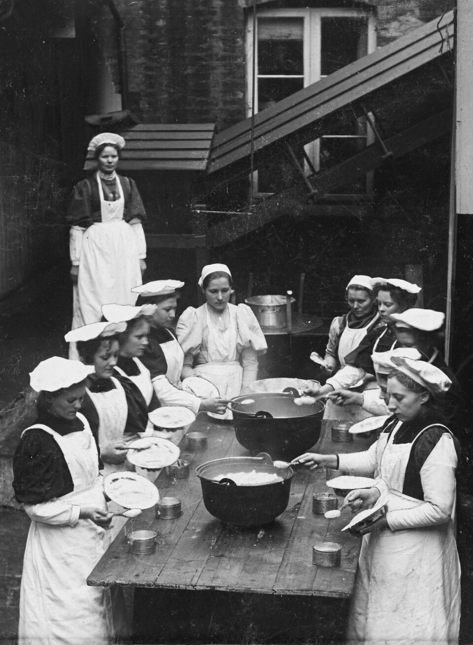 «STAVANGER-EGG»: Enkel fiskebolleproduksjon før århundreskiftet – fiskefarsen på tallerkenene ble formet til boller med en spiseskje, dyppet i kokende vann og så lagt i halvliters hermetikkbokser før gjenlodding og sterilisering. I bakgrunnen står antagelig fabrikkens bestyrerinne.Foto: MUST/Jacobsen