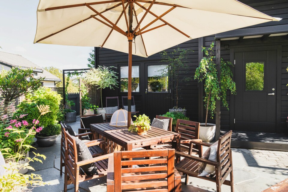 Fremsiden av huset hvor den grå plattingen brytes opp av en raus krukkehage, en koselig spiseplass og parasoll.