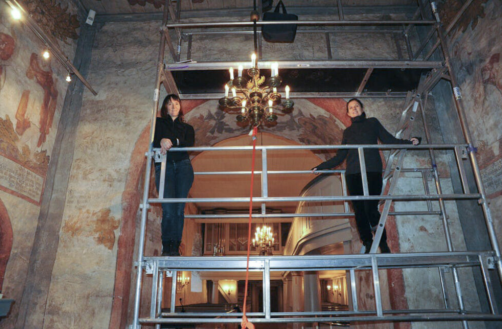 <b>DEMONVEGGEN:</b> Susanne Kaun (t.v.) og Elisabeth Andersen i stillaset foran demonveggen. Demonmaleriene sees som mørke sjatteringer på sidene og over kirkebuen.