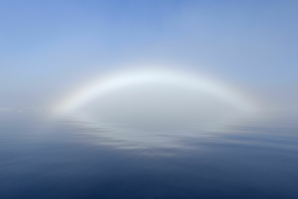 <b>TÅKEBUE:</b> En tåkebue ligner til forveksling på en regnbue, men er ikke helt det samme. Siden vanndråpene i tåke er mindre enn dråpene i skyer, blir fargene mer spredt og blandet så mye at buen blir hvitaktig. En regndråpe kan ha en diameter på cirka én millimeter, mens en gjennomsnittlig tåkedråpe har en diameter på bare cirka 0,01 millimeter. Dette bildet er fra kysten av Svalbard.