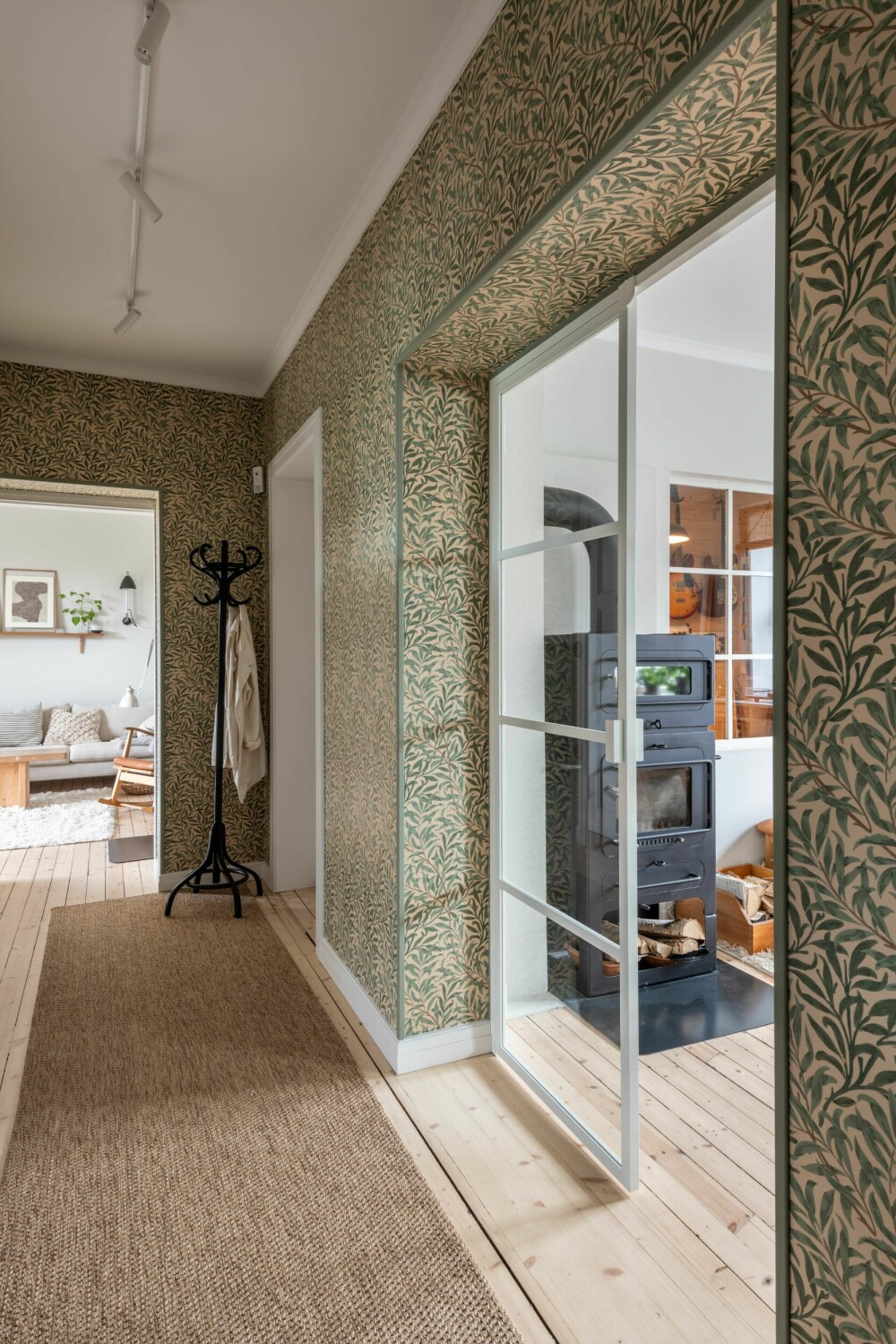 Den lange entreen er kledd med William Morris’ frodige tapet Willow Boughs Green fra 1887. Glassdørene leder inn til allrommet, mens stuen ses helt i enden. 