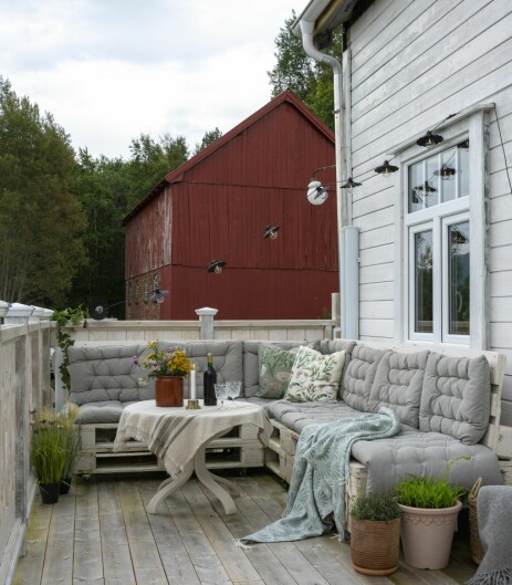 VERANDA: På verandaen har Torunn og Ole Einar laget en koselig sittekrok av paller og nye sitteputer. Bordet har steinplate og tåler å stå ute. Bak ses den store låven som de tenker å innrede til selskapslokale og snekkerverksted en gang i framtiden.
