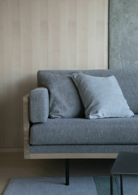 STILRENT DESIGN. Den nette sofaen har både varmt treverk og tidløst tekstil i en grå nyanse, som står godt til materialene brukt på veggen bak.