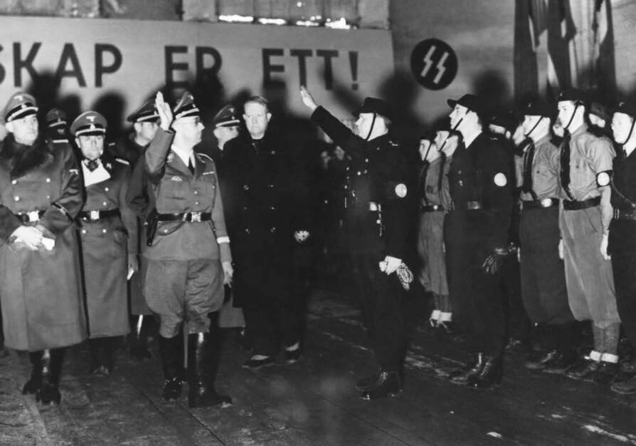 <b>ETT FOLK:</b> Himmler i Norge for å innlemme hird­medlem­mer i rekkene til SS, noe han gjorde to ganger i første halvdel av 1941. Vidkun Quisling til høyre for Himmler. I bakgrun­nen de rune­inspirerte S’ene, et av mange tegn på SS og Himmlers fascinasjon for gammel, germansk kultur.
