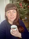 Kvinde med rødt hår og blå øjne holder en kaffekop foran juletræet