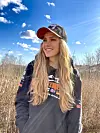 Kvinne i svart Adventure Drive-hettegenser og caps smiler foran siv under blå himmel.