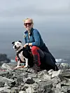 Kvinne i turklær sitter på steiner ved kysten sammen med en hund