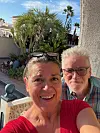To personer tar en selfie på solrik terrasse med palmer og hage bak dem