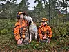 To barn i oransje jaktklær sitter i skogen med en hvit jakthund mellom seg
