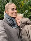 Nicole Ryle i frakke tager selfie og krammer smilende barn i jakke foran grønne buske.