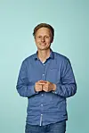 Journalist Anders Eid avbildet smilende i blå skjorte foran turkis bakgrunn.