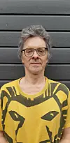 Mann med briller i gul T-skjorte med løvemotiv foran grå trevegg