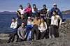 Gruppe mennesker sitter og står på svaberg ved en fjord med fjell bak.