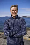 Mann i mørkeblå strikkegenser står med korslagte armer ved havet