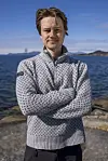 Person i tykk genser står med korslagte armer ved sjøen på solrik dag