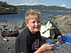 Et barn sitter på svaberg ved fjorden med en kopp i hånden og utsikt over vann og fjell.