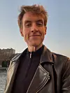 Person i svart skinnjakke tar et selfie ved vannet i skumringslys.