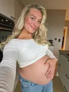 Gravid kvinne i hvit topp og jeans poserer innendørs i et hjem, redigert med Tezza.