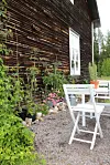 Vit trädgårdsstol vid träbord framför mörk trävägg med tomatplantor och blommor.