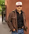 Tobias Sten stående utendørs med brun jakke, svart skjorte, jeans og lys caps med logo.