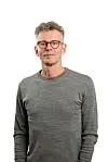 Mann med briller og grå genser står foran hvit bakgrunn.
