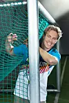 Smilende mann i blå og hvit sportsdrakt bak grønt nett ved en metallstang.