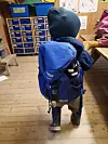 Barn med blå ryggsekk står på et tregulv i et klasserom med fargerike skap og oppslagstavler.