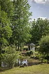 Trädgård med damm, liten vit paviljong och täta gröna träd under en ljus himmel.