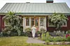 Röd gårdsbyggnad med grön takplåt, blommande urnor vid entrén och en person på gångstigen.
