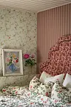 Sovrumshörn med blommig tapet, rosa panel, mönstrad sänggavel, blommiga kuddar och en inramad blomstermålning.