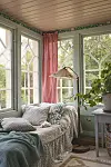 Ljus glasveranda med soffa, golvlampa, gardiner och stora fönster mot grönska.