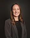 Smilende kvinne i grå blazer og svart topp mot mørk bakgrunn.