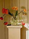 En glasvas med röda och orange blommor står på ett vitt podie mot en ljus vägg.