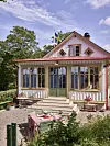 Liten ljus villa med veranda, trädgårdsmöbler och blommande uteplats framför ett trädgårdshus.