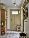 Ljus hall med rutigt golv, beige dörr, träskåp, klädkrokar och en liten pall med rosa kudde.