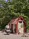 Litet röd-vitt trädgårdshus i en skogsdunge med en skottkärra, en stege och en cykel framför.